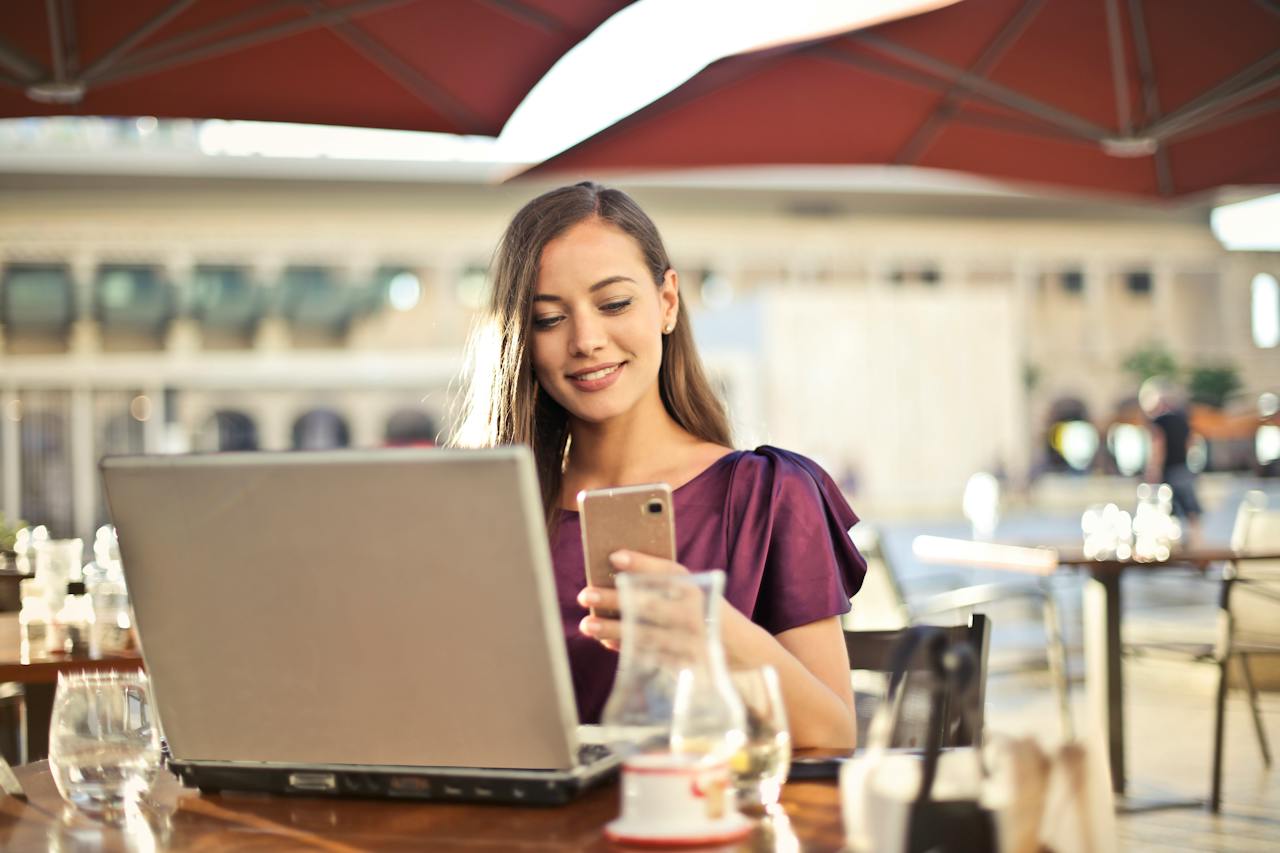 Woman working remotely on a laptop and phone at a sunny cafe