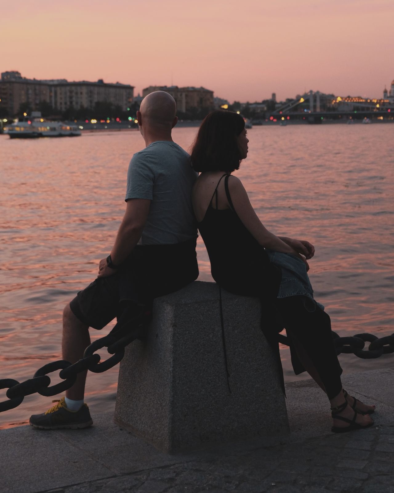 Couple sitting back to back by the water at sunset — reflecting on separate paths forward